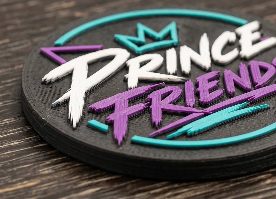 A black 3D-printed coin with the Prince Friends logo raised in white, purple, and teal