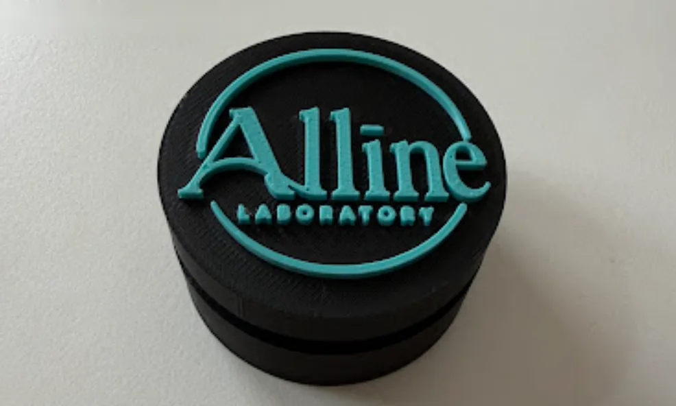A charcoal desk clicker with a raised teal Alline Laboratory logo on top